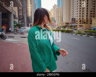 Junge Frau im grünen Anzug geht durch die Straßen von Dubai, Dubai Marina District. Reisekonzept der Vereinigten Arabischen Emirate. Die Vorstellung von erfolgreichem Auswanderer, Umzug in ein anderes Land, Arbeitsvisum Stockfoto