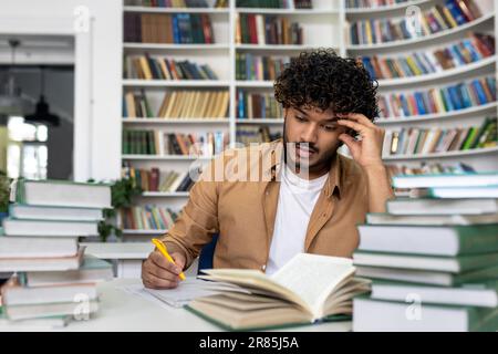Denkender Student, der sich in der akademischen Bibliothek auf die Prüfungen vorbereitet, ein Mann, der überlastet und müde ist, allein zu lernen, ein hispanic, der unter Büchern sitzt und Hausaufgaben macht. Stockfoto
