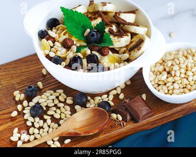 Obstsalat mit Schokolade und Pinienkernen in Joghurt auf einem weißen Teller. Blick von oben. Gesundes nahrhaftes Frühstück mit appetitlichen Cornflakes Salat mit Blaubeeren und Minze. Stockfoto