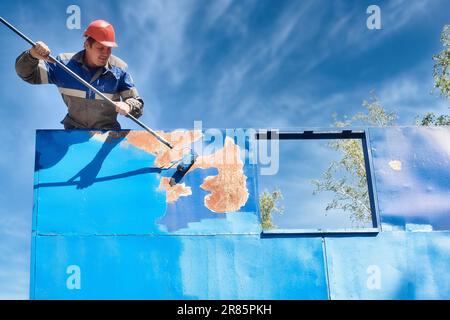 Ein industrieller Profi-Maler mit Schutzhelm und Arbeitskleidung malt an einem Sommertag eine Metalloberfläche mit einer Rolle. Stockfoto