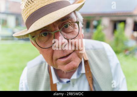 Ein fröhlicher und leicht betrunkener Senior Mann, der einen Strohhut, ein Hemd, eine Brille und eine Schürze trägt, sieht merkwürdigerweise in die Kamera. Das Foto wurde gemacht Stockfoto
