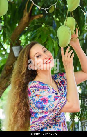 Blonde, schöne Frau, die Mangofrüchte vom Baum anrührt Stockfoto