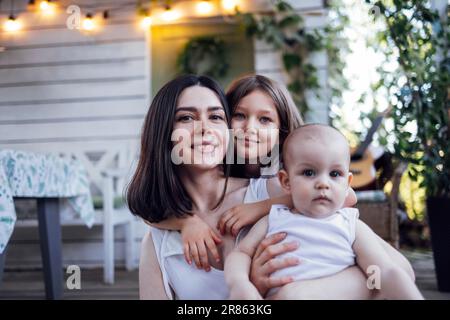 Wunderschöne, lächelnde brünette mutter mit zwei süßen kleinen Kindern. Eine glückliche Familie umarmt sich auf der Terrasse des Holzhauses im Vintage-Stil. Retro-Interieur mit Tisch, Stockfoto