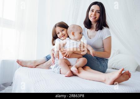 Wunderschöne, lächelnde brünette mutter mit zwei süßen kleinen Kindern. Hübsches Schulmädchen umarmt ihren kleinen Bruder. Charmante junge Frau mit ihren Kindern sitzt auf Be Stockfoto