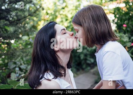 Wunderschöne brünette Frau, die ihre kleine Tochter auf der Nase küsst, während sie im Park spaziert. Glückliche mutter und Kind umarmen sich und haben Spaß zusammen im Garten. Ca Stockfoto