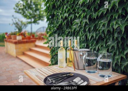 Drei Flaschen leichten Weins, Eiskübel, Glaskannen mit Wasser, Gabeln und Löffel auf einem Tablett auf einem Holztisch. Vor der Gartenparty. Die Wand ist mit Efeu bedeckt. Stockfoto