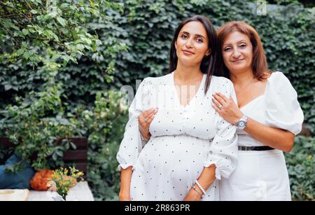 Die attraktive, schwangere Brünette lässt ihre mutter im Garten im Freien kuscheln. Süße lächelnde Frauen in eleganten weißen Kleidern umarmen sich im Park. Eine Frau erwartet Stockfoto