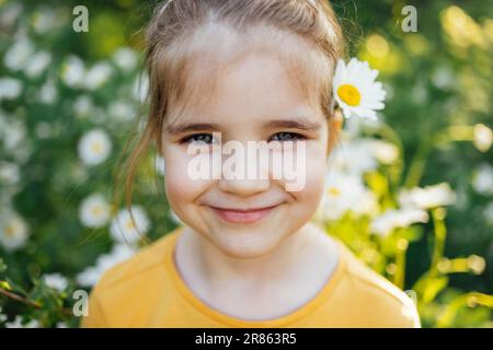 Kinderporträt eines süßen kleinen blauäugigen Mädchens mit Kamillenblume hinter dem Ohr im Park oder Garten. Süßes Mädchen lächelt und schaut draußen in die Kamera Stockfoto