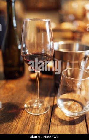 Nahaufnahme eines stillen Lebens eines eleganten Bechers mit Rotwein und einem Glas Wasser auf einem strukturierten Holztisch. Ein Eiskübel und eine dunkle Glasflasche Stockfoto