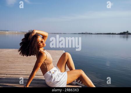 Das wunderschöne gemischte Rennmädchen sitzt auf einem hölzernen Pier und genießt die Natur. Eine charmante Studentin in beigefarbenem Oberteil und weißen Shorts hat ihre Augen geschlossen Stockfoto