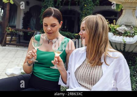 Zwei attraktive Freundinnen in lässiger Kleidung sitzen im Open-Air-Café. Junge Frauen trinken Champagner und Klappergläser. Charmante Mädchen plaudern und so Stockfoto