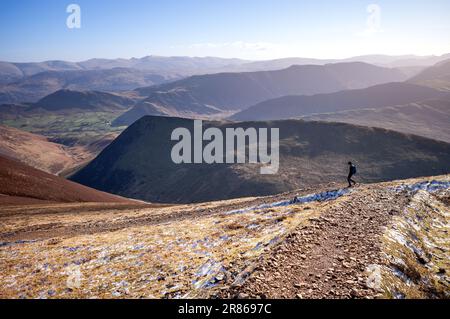 Eine Wanderin und ihr Hund steigen vom Gipfel des Sail in Richtung Causey Pike auf einem felsigen Pfad hinab, mit ARD Crags in der Ferne im Winter im Engl Stockfoto