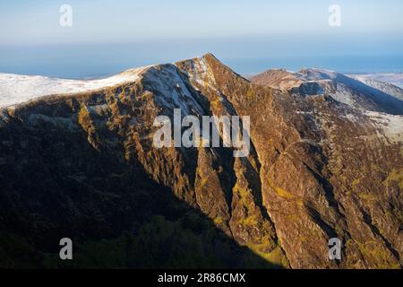 Ein entfernter Wanderer auf dem Gipfel des Hopegill Head mit den steilen Klippen von Hobcarton Crag im Winter im englischen Lake District, Großbritannien. Stockfoto