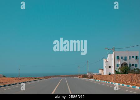Breite, leere zweispurige Straße in MIRLEFT, Marokko. Flaches weißes Gebäude mit blauen Fenstern an der Seite. Küstenstadt in Afrika. Lokaler Immobilienhintergrund. Stockfoto