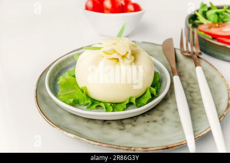 Geschnittene italienische Käseburrata wird auf einem runden Teller mit Basilikum und Tomaten mit einer Gabel und einem Messer auf dem Hintergrund eines alten Baumes serviert. Stockfoto