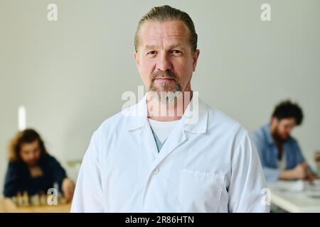 Selbstbewusster, reifer Psychiater im Laborkittel steht vor der Kamera gegen zwei Patienten der psychiatrischen Klinik und schaut Sie an Stockfoto