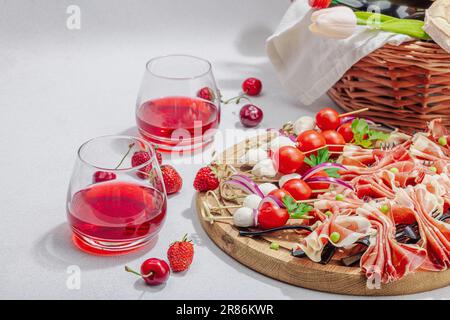 Picknick-Food-Konzept. Korb mit Wein und Baguette, Snacks auf Spießen. Fleisch, Käse, Gemüse, Obst und Blumenstrauß. Hartes Licht, dunkle s Stockfoto