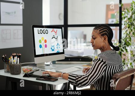 Seitenansicht einer jungen, seriösen afroamerikanischen Webdesignerin, die am Arbeitsplatz vor dem Computermonitor sitzt und ein Tablet benutzt Stockfoto