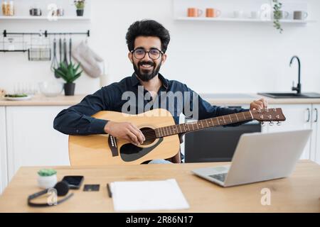 Porträt eines gutaussehenden bärtigen Erwachsenen, der mit einer Gitarre mit sechs Bändern posiert, während er zu Hause einen angenehmen Zeitvertreib genießt. Lächelnder indianer in einer Brille lernt Instrument spielen, indem er Online-Unterricht auf einem Laptop nimmt. Stockfoto