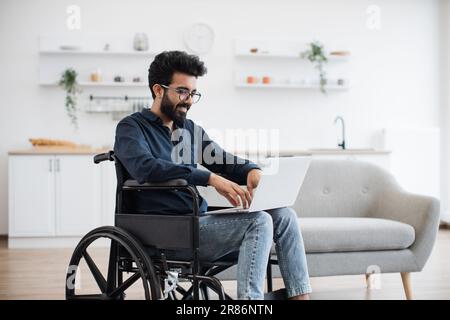Ein fröhlicher indianer im Rollstuhl hielt sein Notebook auf Knien, während er mitten in einer hellen, offenen Küche blieb. Glücklicher junger Erwachsener, der im Internet surft, während er sich nach einem Unfall zu Hause erholt. Stockfoto