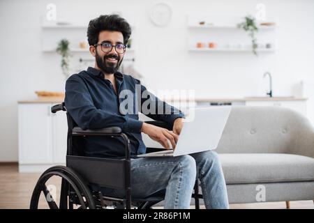 Ein fröhlicher indianer im Rollstuhl hielt sein Notebook auf Knien, während er mitten in einer hellen, offenen Küche blieb. Glücklicher junger Erwachsener, der im Internet surft, während er sich nach einem Unfall zu Hause erholt. Stockfoto