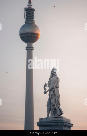 Eine beleuchtete Statue einer Person steht in der Nähe eines hohen Turms bei Sonnenuntergang, mit dem Fernsehturm im Hintergrund Stockfoto