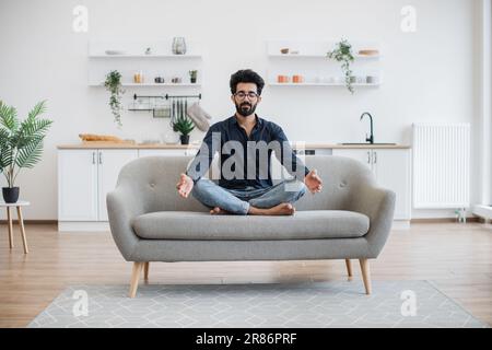 Blick auf die gesamte Länge einer erwachsenen Person, die Yoga macht, während sie mit überkreuzten Beinen auf der Couch im Esszimmer sitzt. barfußindianer in Brille entspannen mit geschlossenen Augen nach einem Arbeitstag zu Hause. Stockfoto