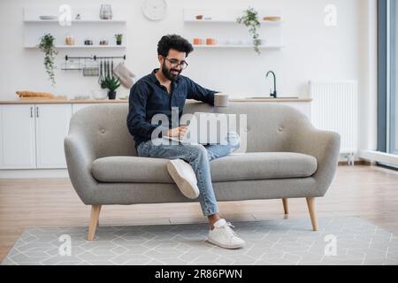 Blick über den gesamten Fußweg des gutaussehenden bärtigen Mannes mit einer Tasse Tee in der Hand, der ein modernes Notebook benutzt, während er sich zu Hause auf dem Sofa ausruht. Junger arabier mit Brille, der in der Freizeit drinnen im Internet surft. Stockfoto