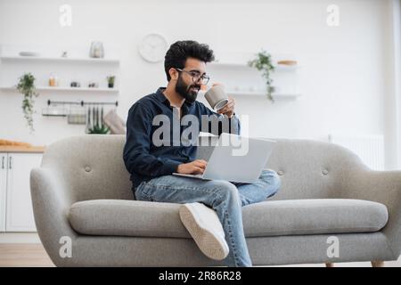 Gutaussehender Mann mit Bart und einer Tasse Tee in der Hand, der ein modernes Notebook benutzt, während er sich zu Hause auf dem Sofa ausruht. Junger arabier mit Brille, der in der Freizeit drinnen im Internet surft. Stockfoto