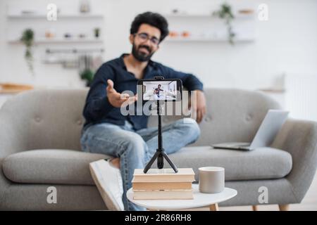 Positiver indischer Einflussnehmer mit schwarzem Bart, der Videos auf einem modernen Smartphone aufnimmt, während er mit einem Laptop auf dem Sofa sitzt. Glücklicher junger Mann, der seinen Erfolg online mit Abonnenten teilt. Stockfoto