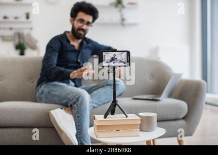 Positiver indischer Einflussnehmer mit schwarzem Bart, der Videos auf einem modernen Smartphone aufnimmt, während er mit einem Laptop auf dem Sofa sitzt. Glücklicher junger Mann, der seinen Erfolg online mit Abonnenten teilt. Stockfoto