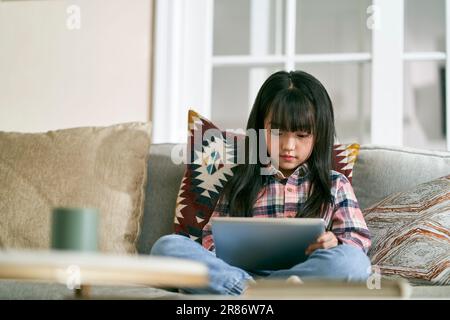 7-jähriges kleines asiatisches Mädchen, das zu Hause auf dem Familientisch sitzt und einen digitalen Tablet-Computer benutzt Stockfoto