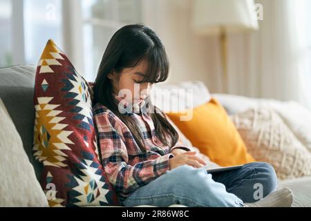 7-jähriges kleines asiatisches Mädchen, das zu Hause auf dem Familientisch sitzt und einen digitalen Tablet-Computer benutzt Stockfoto