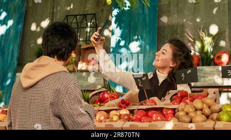 Junger Landwirt, der mit einem normalen Kunden ein Video auf einem Smartphone aufnimmt, um kleine landwirtschaftliche Betriebe in sozialen Medien zu fördern. Weibliche Verkäuferin filmt mit Handy, amüsiert sich mit dem Kunden. Stockfoto