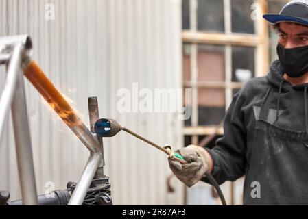 Junger spanischer Mann, der mit einer Lötlampe die Farbe eines orangefarbenen Fahrradrahmens entfernt, der gerade in seiner Werkstatt renoviert wird. Stockfoto