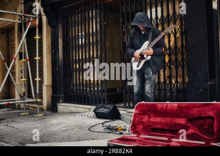 Ein unbekannter, mit Kapuze besetzter Straßenmusiker in einer Fußgängerzone im Stadtzentrum, der eine elektrische Blues-Gitarre mit Verstärker und Pedalen spielt Stockfoto