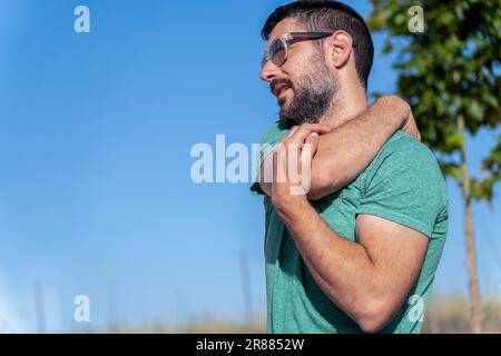 Profilansicht eines attraktiven bärtigen Mannes mit einer Sonnenbrille, die seine Schultern auf einem Feld mit blauem Himmel im Hintergrund dehnt Stockfoto