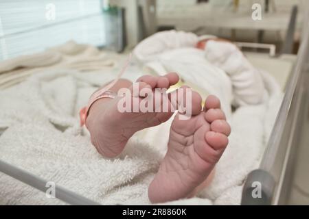 Nahaufnahme eines winzigen Paar Füße und Zehen eines Neugeborenen, das in eine Decke gewickelt ist und ein Identitätsarmband aus Plastik trägt, im Kreißsaal des hospit Stockfoto