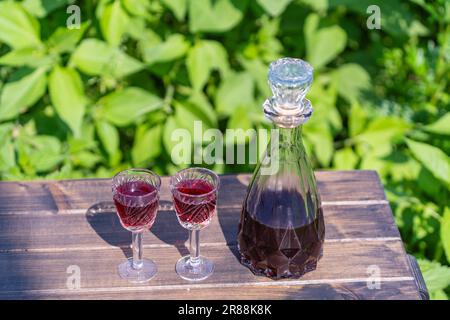 Hausgemachter Kirschbranntwein in Gläsern und in einer Glasflasche auf einem Holztisch in einem Sommergarten, Nahaufnahme Stockfoto