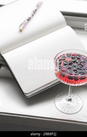 Ein offenes Notizbuch mit leeren Seiten und ein Glas roter Cocktail mit Blaubeeren auf einer Fensterbank Stockfoto