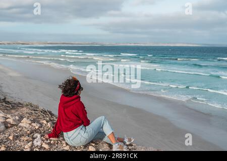 Eine Frau mit kurzen Haaren und rotem Kapuzenpulli, die an einem felsigen Strand mit Blick auf das Meer sitzt Stockfoto