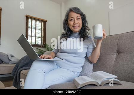 Angenehmes Gespräch. Schöne ältere Frau, die auf ihrem Laptop SMS schreibt und glücklich lächelt, während sie Kaffee trinkt und auf der Couch im Wohnzimmer sitzt Stockfoto
