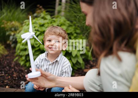 Portriat des süßen Jungen hält Windturbine, während Vater sein Elektroauto auf der Straße auflädt. Stockfoto