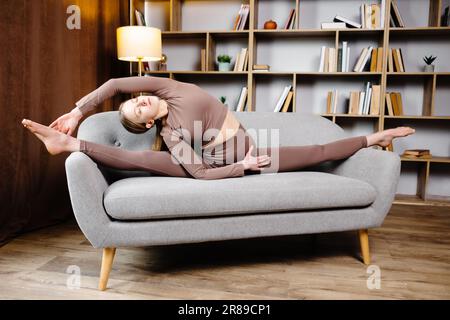 Ein süßes, sportliches Mädchen sitzt in einer gespaltenen Pose auf der Couch und streckt ihre Hand bis zum Bein. Stockfoto