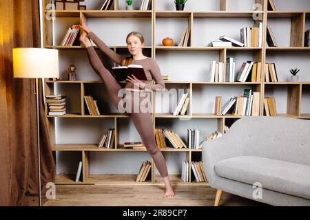 Junge Frau in Sportbekleidung, die sich Beine streckt, Spaltungen macht, in der Bibliothek steht und Buch liest. Die Frau beim täglichen Workout macht zu Hause Yoga-Training. Stockfoto