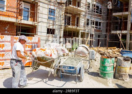 Arbeiter mit Schutzhandschuhen hält den Griff der Schubkarre voll mit zerbrochenem Zellophan, das der Kran auf die Baustelle legt. Im Hintergrund ist unf Stockfoto