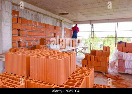 Anhäufung roter Keramikblöcke auf Holzplattform auf der Baustelle. Im Hintergrund baut der Arbeiter Trennwand mit Blöcken und Mörtel. Stockfoto