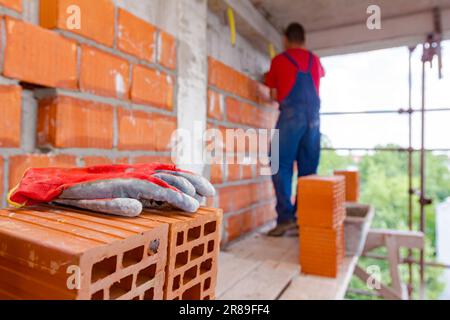 Arbeiterschutzhandschuh auf roten Keramikblöcken auf Holzplattform auf der Baustelle. Im Hintergrund baut der Arbeiter eine Trennwand mit Blöcken A. Stockfoto