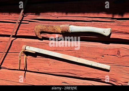 Zwei alte landwirtschaftliche Hacke-Werkzeuge hängen an der Außenseite der traditionellen Holzwand, die mit roter Ockerfarbe gestrichen ist. Freilichtmuseum Kreivinmaki, Salo, FI. Stockfoto