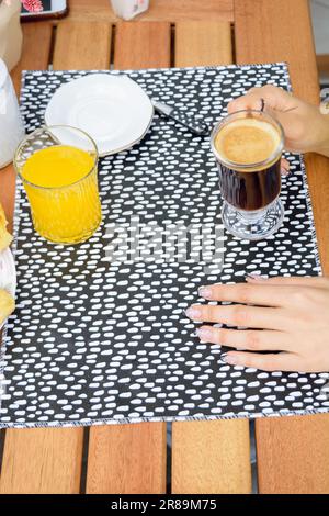 Vertikales Bild von nicht erkennbaren weiblichen Händen, die eine Tasse Kaffee auf dem Tisch halten, außerhalb der Cafeteria während des Frühstücks, mit Kopierbereich. Stockfoto
